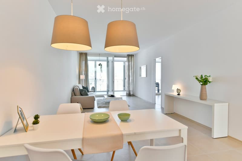 Bright living and dining area with white walls, large windows, and a balcony. Features a sofa, coffee table, dining table with chairs, and decorative items.