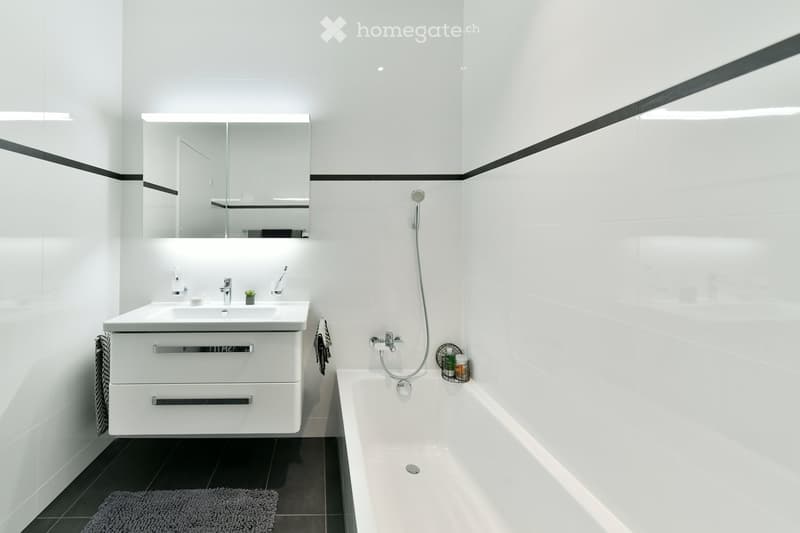 Modern bathroom with white tiles, a large mirror, a sink with a cabinet, a bathtub, and a showerhead. The space includes a towel, a bathrobe, and a small plant on the sink.