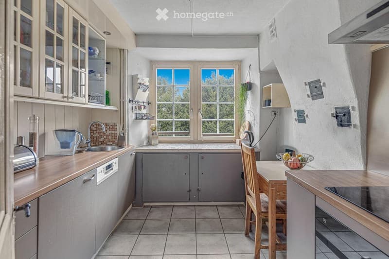 Bright kitchen with wooden countertops, stainless steel sink, dishwasher, and wooden dining area with a fruit bowl. Large windows provide natural light and a view of greenery.