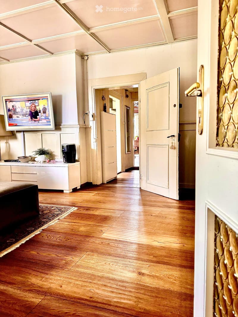 Wooden floor, open door leading to another room, mounted TV, white cabinet with decorative items, carpet, open hallway