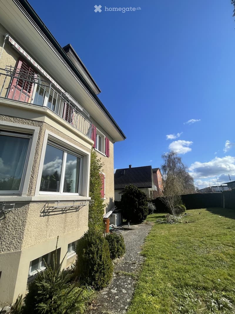Two-story house with beige siding, large windows, and a balcony on the upper level. The house features red shutters and is surrounded by a well-maintained garden with a gravel pathway.