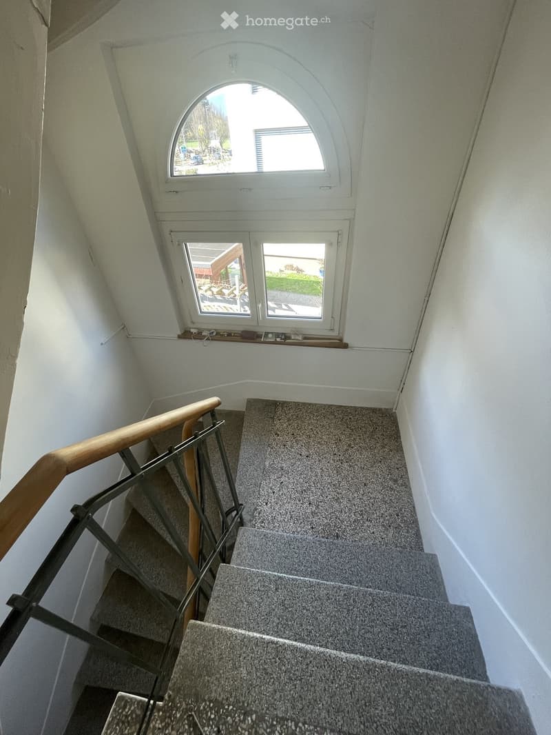 Staircase with grey carpeted steps, wooden handrail, and metal balustrade. The walls are white, and there is a window at the top of the stairs.