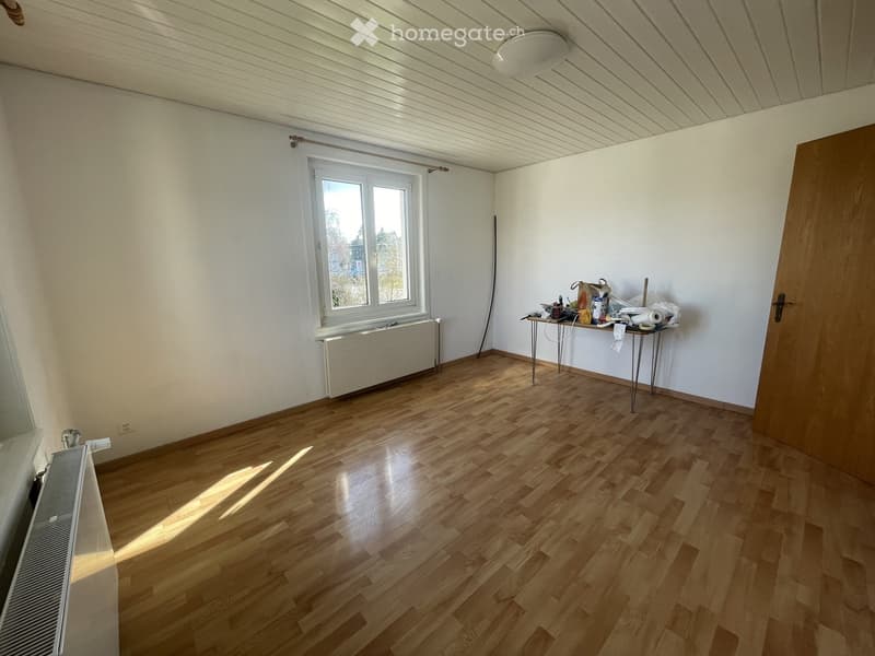 Empty room with wooden flooring, white walls, a window with a radiator below, and a small table with miscellaneous items on it.