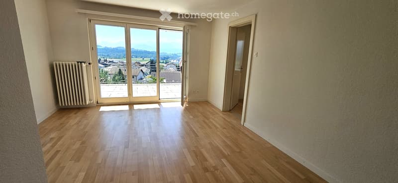 Empty room with wooden flooring, large sliding glass door leading to a balcony, radiator on the left side, and a doorway on the right.