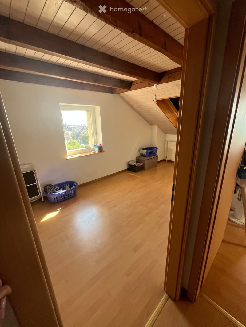 An attic room with wooden floors, exposed wooden beams, and a skylight window. The space is empty, with only a few storage items visible near the window and a small folding chair in the corner.