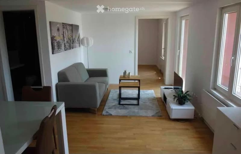 Modern living room with light wooden flooring, grey sofa, coffee table, small side table, and a plant on a white console. The room features large windows and a hallway leading to another area.