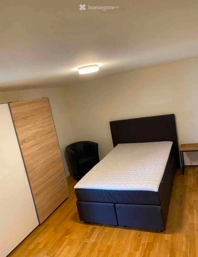 A small, furnished bedroom with a bed, a chair, and a wardrobe. The room has wooden flooring and white walls.