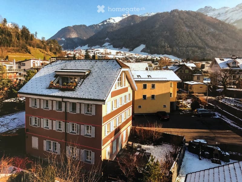 Multi-story house, snow-covered roof, multiple windows, several neighboring houses, mountainous background, parked cars, surrounded by trees and snow-covered ground.