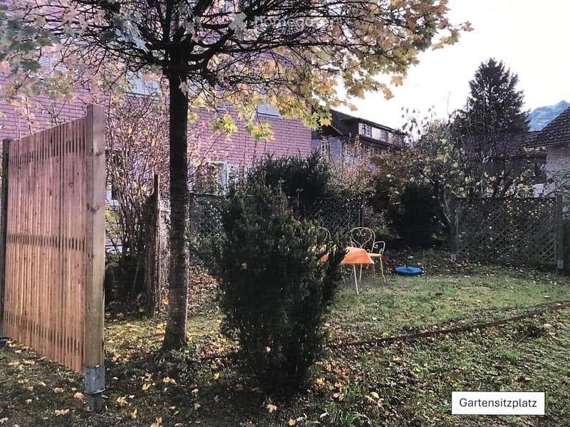 garden with lawn, shrubs, wooden fence, tables and chairs, buildings in background