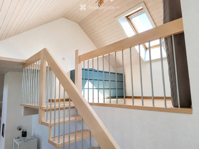 Modern wooden staircase with metal balustrade, leading to an upper level with a skylight window and a view of a room below.