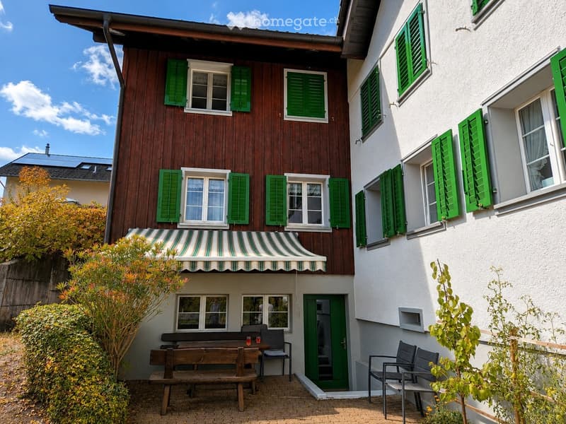 Two-story house with green shutters, white walls, and a striped awning over the entrance. The house features a small patio area with a wooden bench, chairs, and a table.