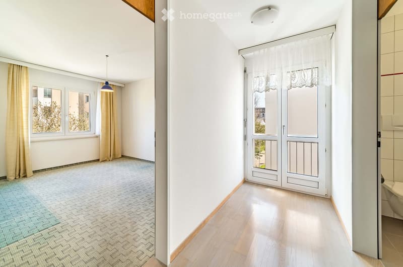 Empty room with white walls, wooden flooring, and large windows with curtains. The room is unfurnished and has a neutral color scheme.