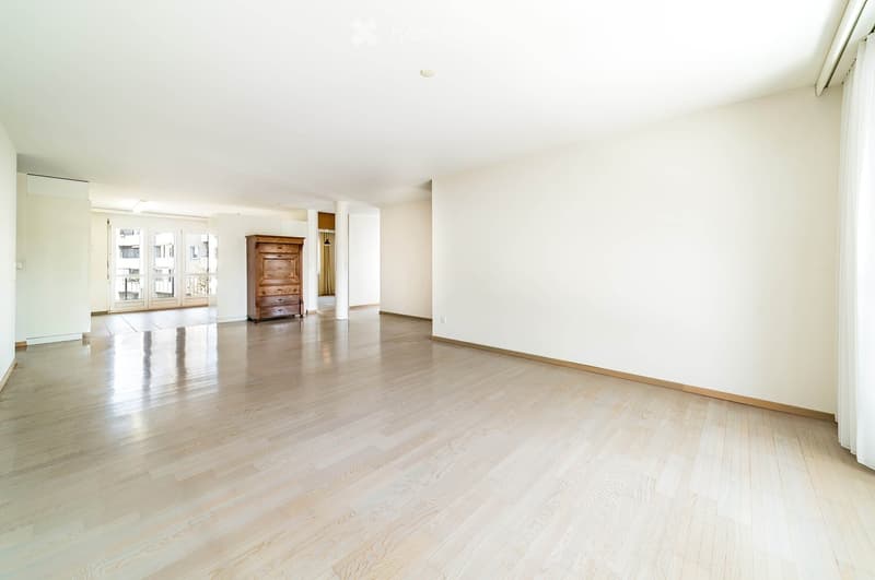Bright, spacious room with light wooden flooring and white walls. A single dresser is placed against one wall, and large glass doors lead to a balcony area.
