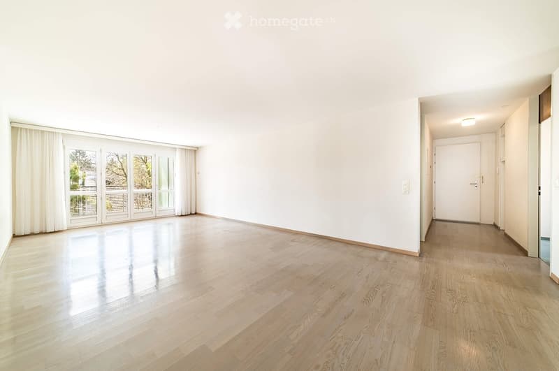 Empty room with light wooden flooring, white walls, large glass doors with curtains, and a hallway leading to another room.