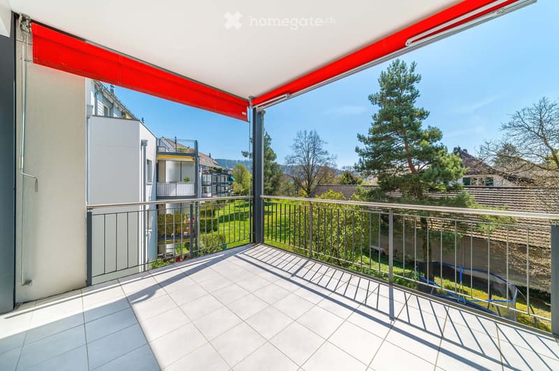 Balcony with tiled floor, metal railing, retractable awning, overlooking a green area with trees and neighboring buildings.