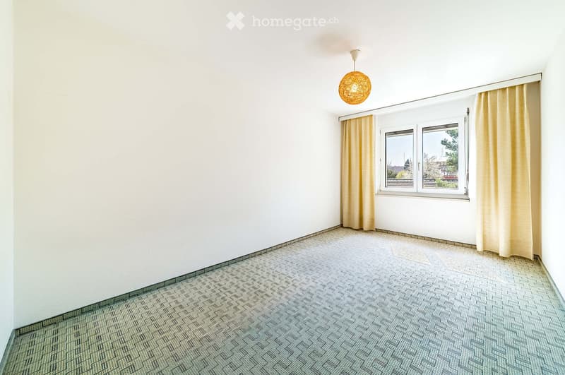 Empty room with white walls, patterned carpet, and a window with beige curtains. A single hanging light fixture is visible.