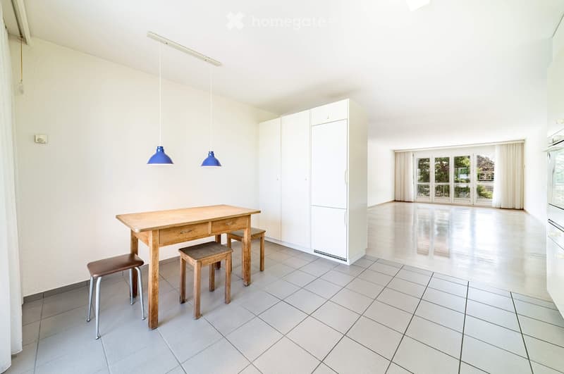 Bright, minimalist room with white walls, tiled floor, and large windows. Features a wooden table with two stools, white cabinetry, and blue pendant lights.