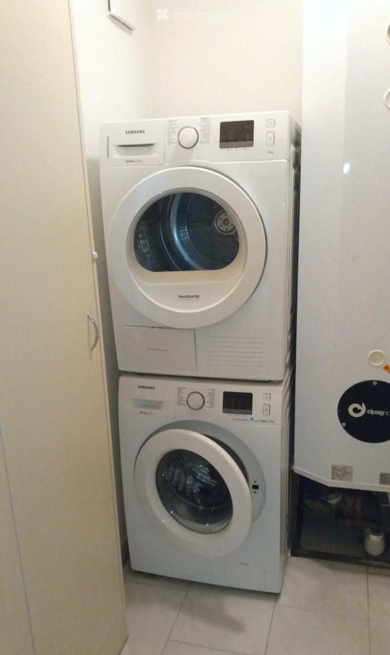 A compact bathroom with a stacked Samsung washing machine and dryer unit, white tiled floor, and light-colored walls.