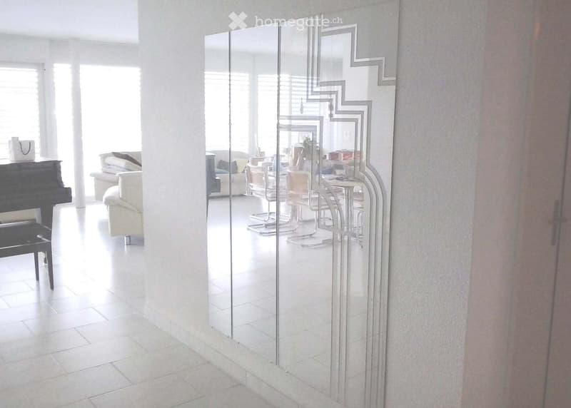 A bright hallway with white tiled flooring, large mirrored wall panels, and a view into an adjacent living area with a sofa, chairs, and a dining table.