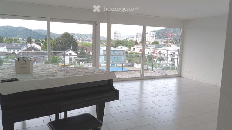 Bright room with large windows offering a view of the city and surrounding hills. Features a grand piano with a mattress placed on top, a piano bench, and a shopping bag on the floor.