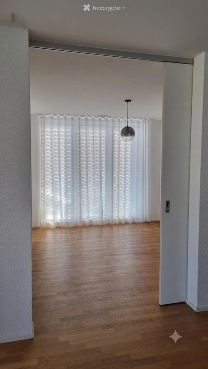 Empty room with wooden flooring, white walls, and large windows covered with white curtains. A single pendant light hangs from the ceiling.