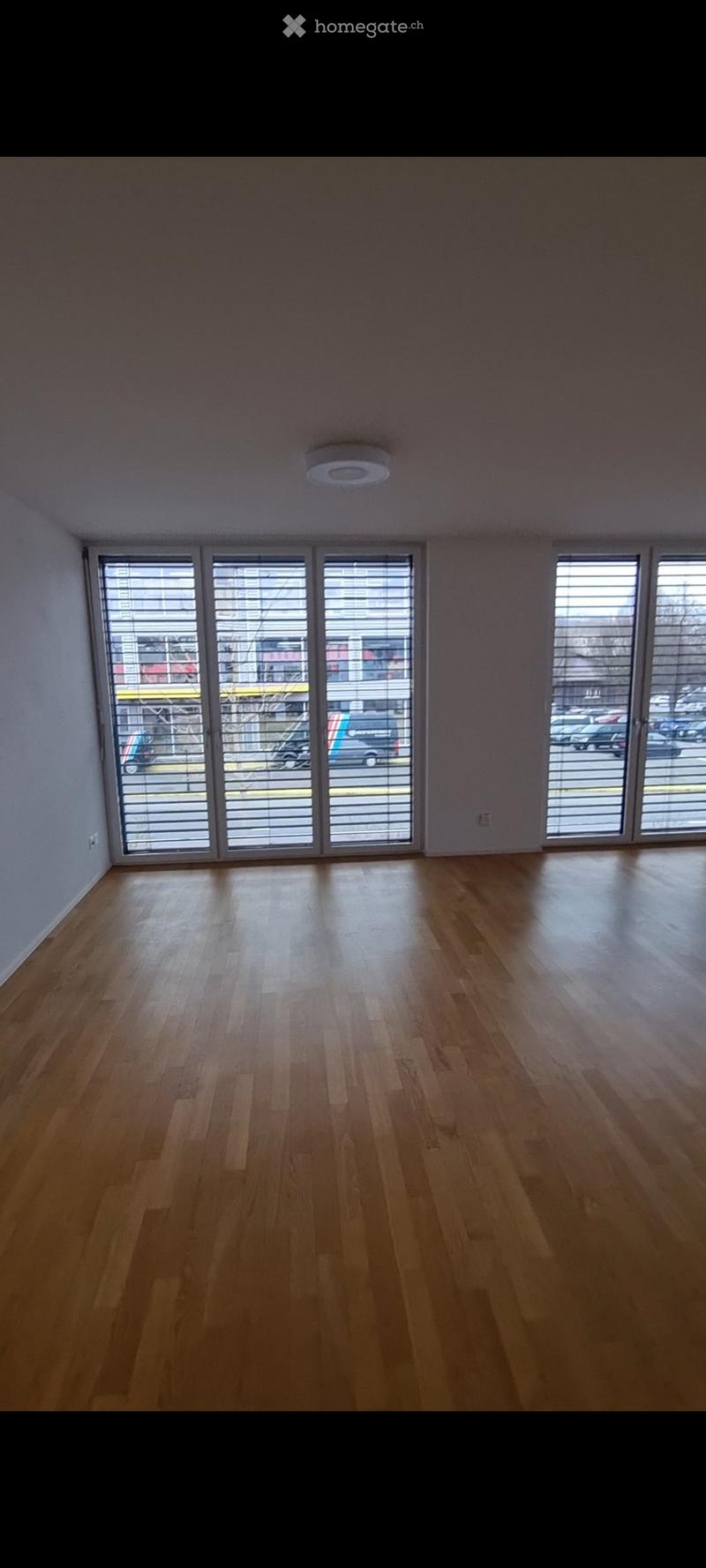 Empty room with wooden flooring, large windows with blinds, and white walls.