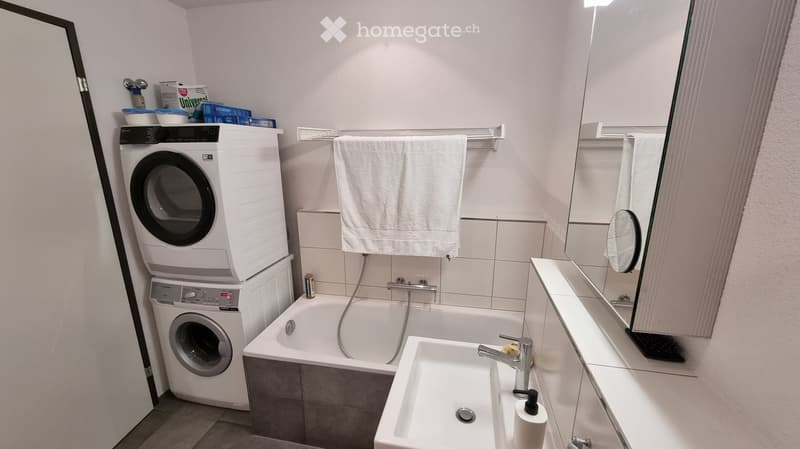 Modern bathroom with a bathtub, sink, washing machine, and dryer stacked together, a large mirror, and a towel hanging on a rack.