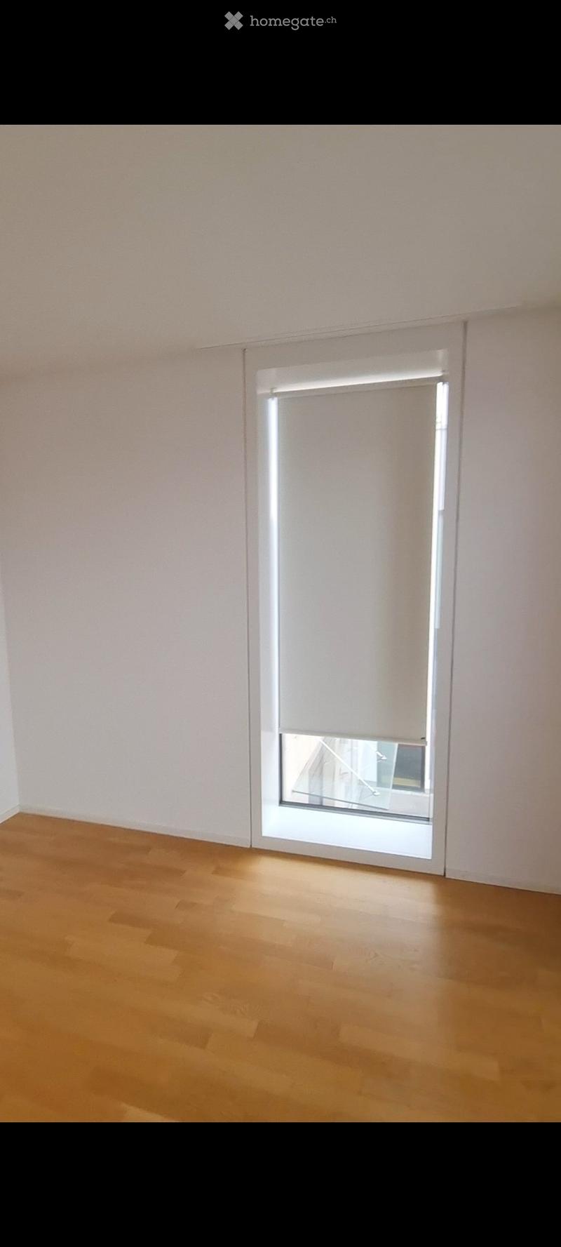 Empty room with light wooden flooring and white walls, featuring a large window with blinds.