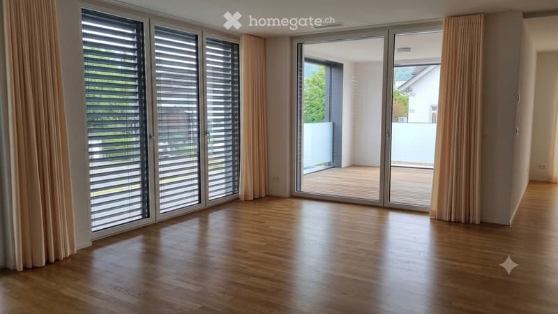 Empty room with wooden flooring, large sliding glass doors with blinds, and light-colored curtains. The space is unfurnished and appears to be a living or dining area.