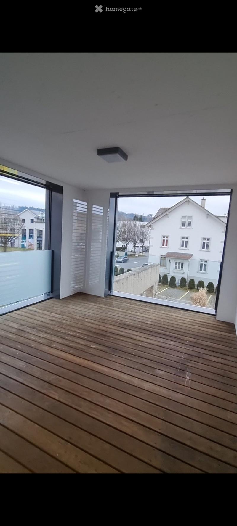 Empty wooden balcony with large glass doors opening to a street view, modern lighting fixture on the ceiling.