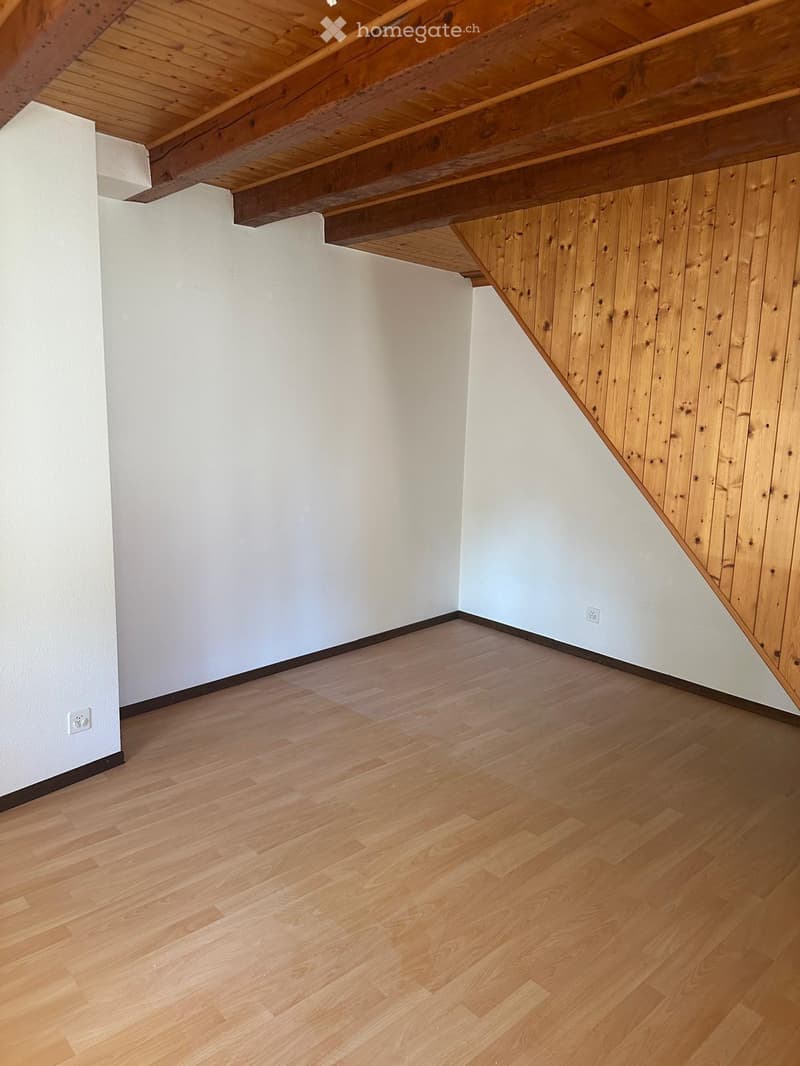 Empty room with light wooden flooring, white walls, and exposed wooden beams on the ceiling.