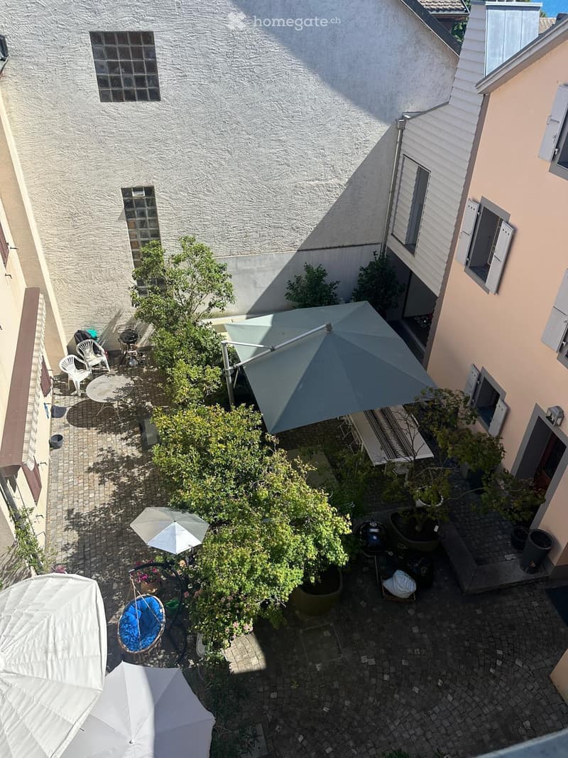 Courtyard with a table, chairs, umbrellas, potted plants, and a grill, surrounded by buildings.