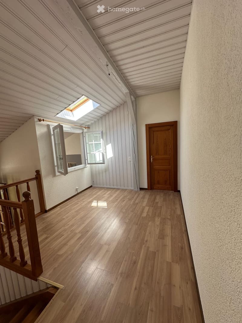 Spacious hallway with wooden flooring, white walls, and a wooden staircase railing on the left. A wooden door is visible on the right, and a skylight brings in natural light.