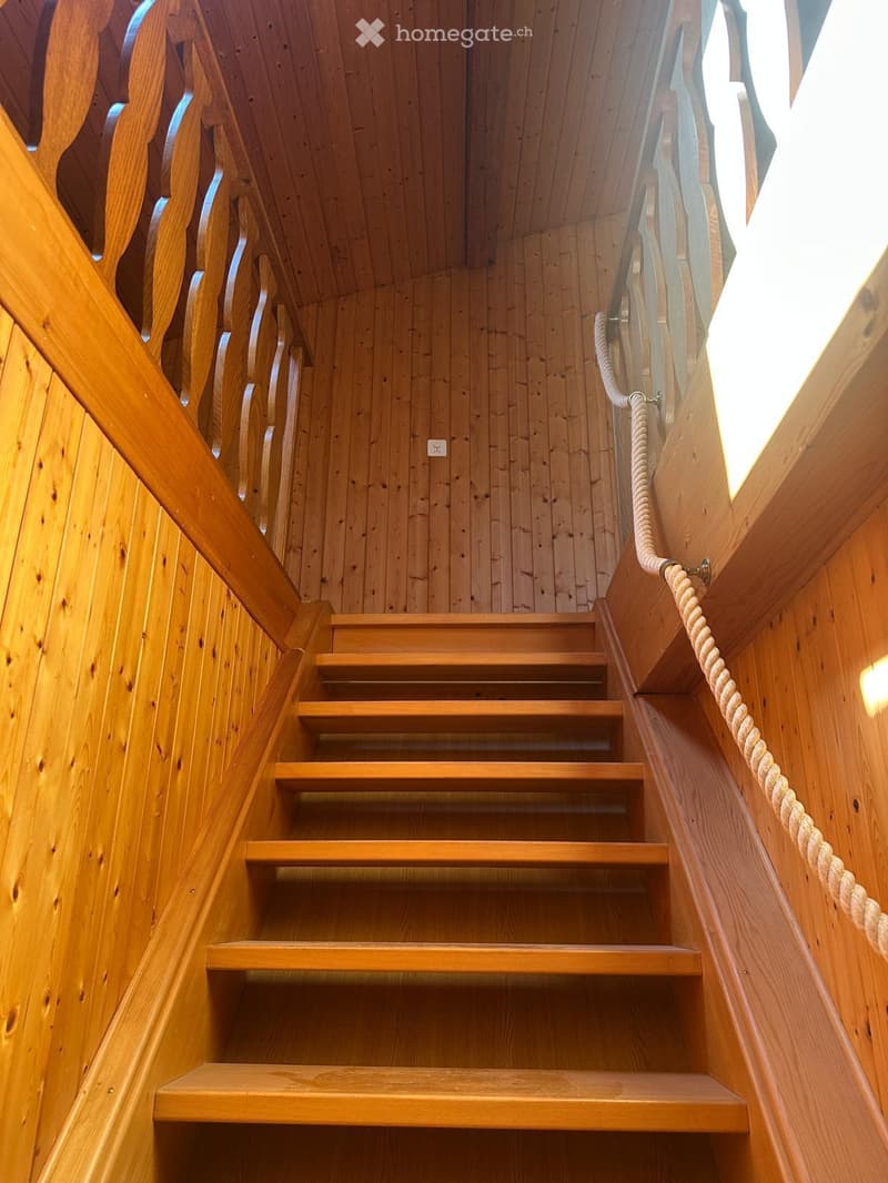 Wooden staircase with rope handrail, wooden walls, and ceiling, leading upwards.