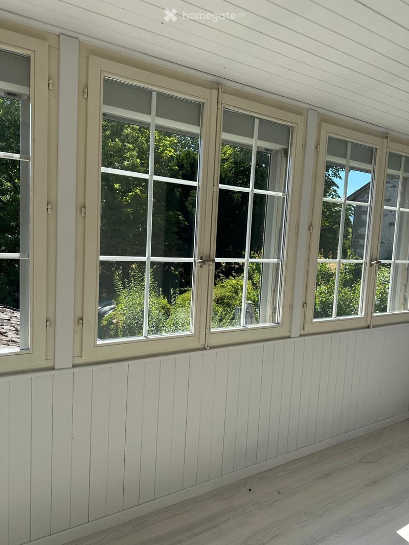 Balcony with wooden paneling, large windows, and a view of greenery outside.