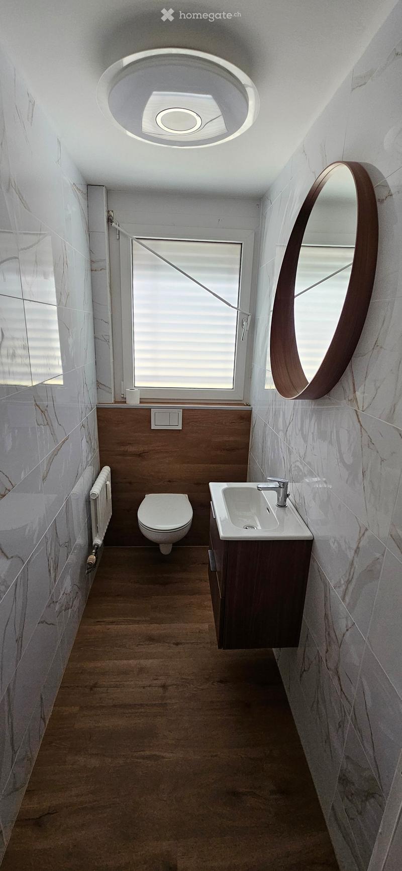 Modern bathroom with marble walls, wooden floor, toilet, sink, and a round mirror with wooden frame.
