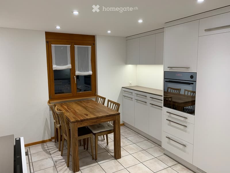 Modern kitchen with white cabinets, a wooden dining table, and six chairs. Features a built-in oven and a large window with wooden frames.