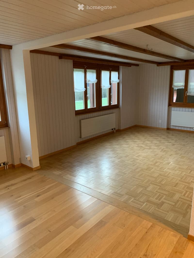 Empty room with wooden flooring, exposed wooden beams on the ceiling, and large windows with white curtains. The walls are paneled in light wood, and there are radiators along the walls.