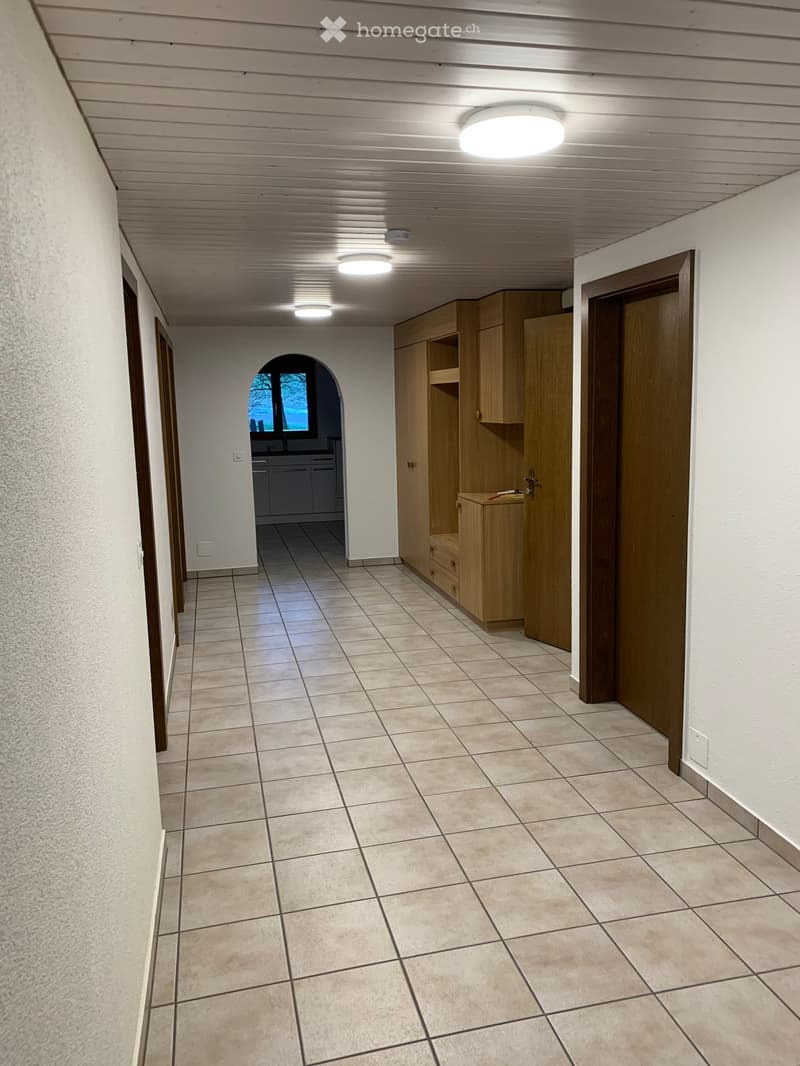 Empty hallway with tiled floor, wooden cabinet, and recessed lighting. An arched window at the end provides natural light.