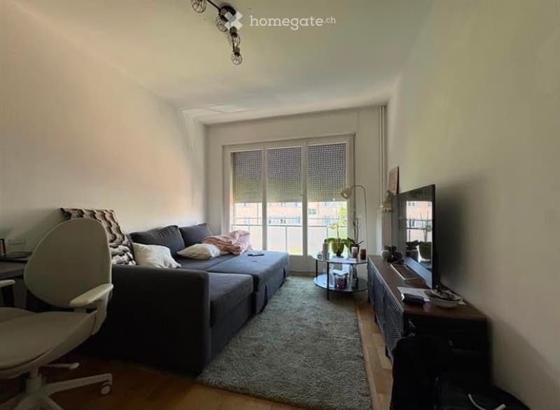 Modern living room with a grey sectional sofa, coffee table, TV stand, floor lamp, and a small round table near the balcony. The room features large windows with blinds and a cozy rug on the floor.