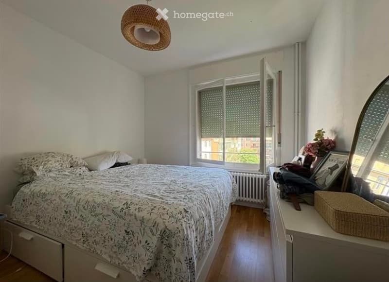 Cozy bedroom with a large bed, nightstand, mirror, wicker basket, and a window with blinds. The room features wooden flooring and white walls.
