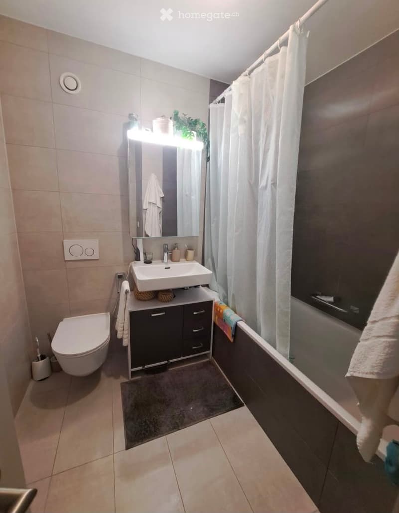 Modern bathroom with a white sink, mirror with built-in lighting, toilet, bathtub with a white curtain, and a small plant on the sink counter.