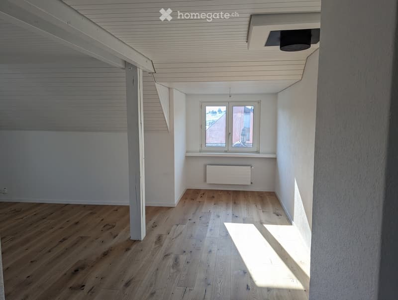 Empty room with wooden flooring, white walls, and a window with a view of the outside. The ceiling has exposed wooden beams and a modern light fixture.