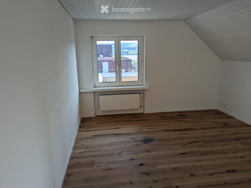 Empty room with wooden flooring, white walls, and a large window offering a view of the outside. A radiator is visible below the window.