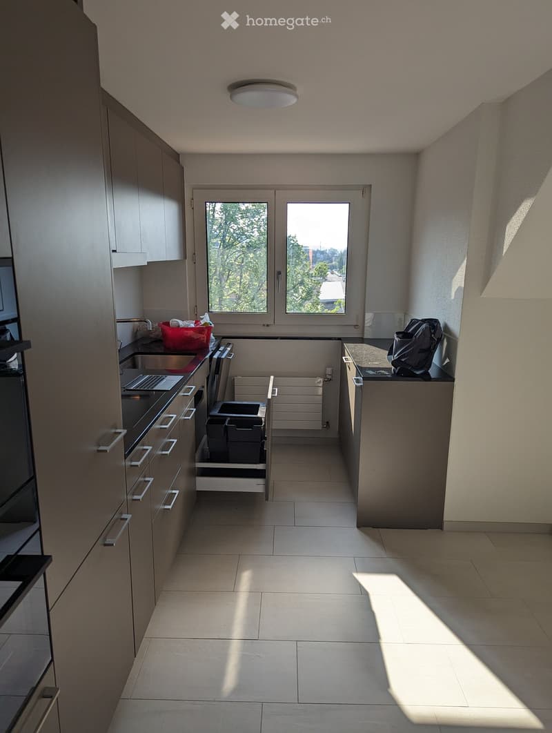 Modern kitchen with light grey cabinets, tiled floor, and large windows letting in natural light. Features a sink, laptop, backpack, and trash bin.