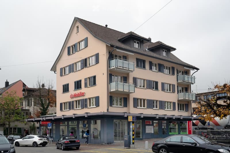 Multi-story building with beige facade, multiple windows with shutters, and balconies on the upper floors.
