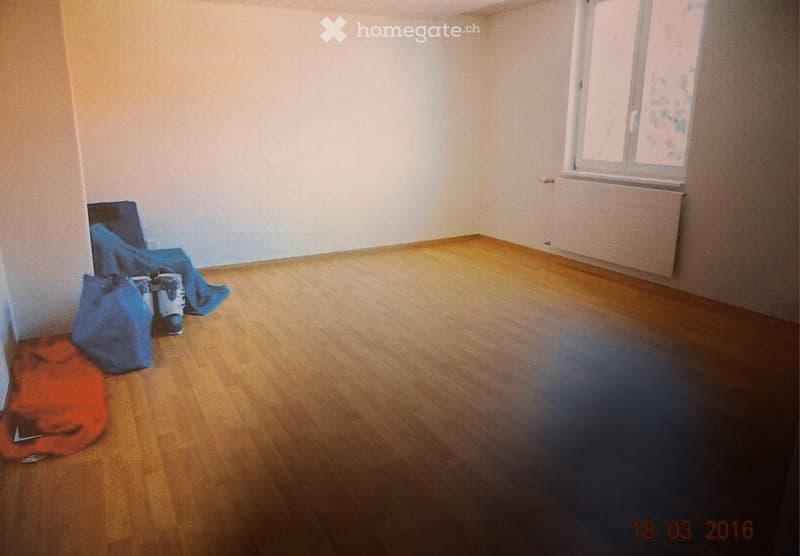 Empty room with wooden flooring, white walls, and a single window. There are a few bags and a pair of shoes placed near the wall.