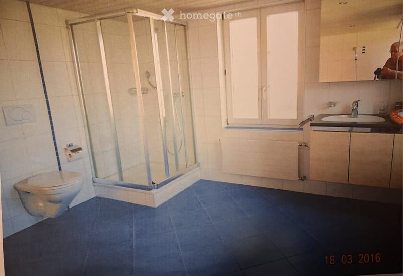 Modern bathroom with blue tiled floor, white tiled walls, a glass-enclosed shower, a toilet, a sink with a cabinet, and a window for natural light.