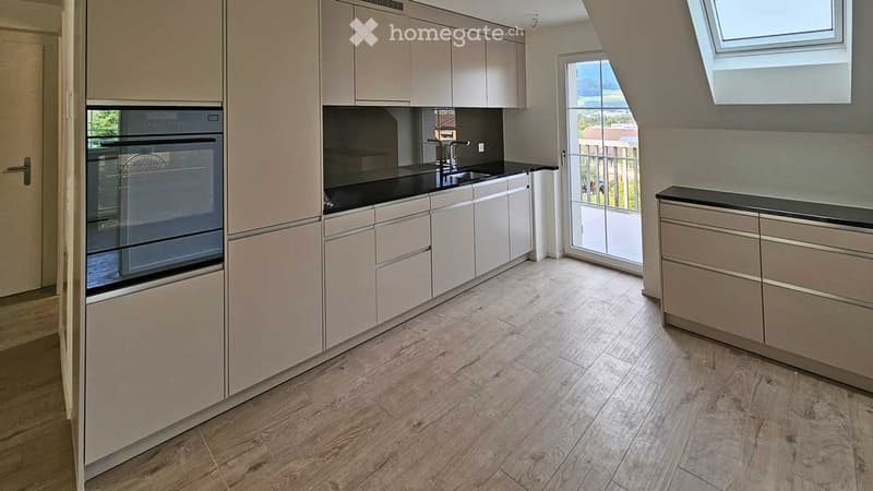Modern kitchen with light-colored cabinets, black countertops, wooden flooring, and a view of a balcony through large windows.
