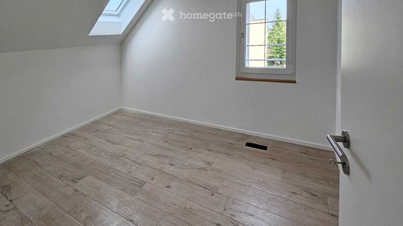 Empty room with light wooden flooring, white walls, and a window with a view of greenery. The room features a skylight and a small vent on the floor.